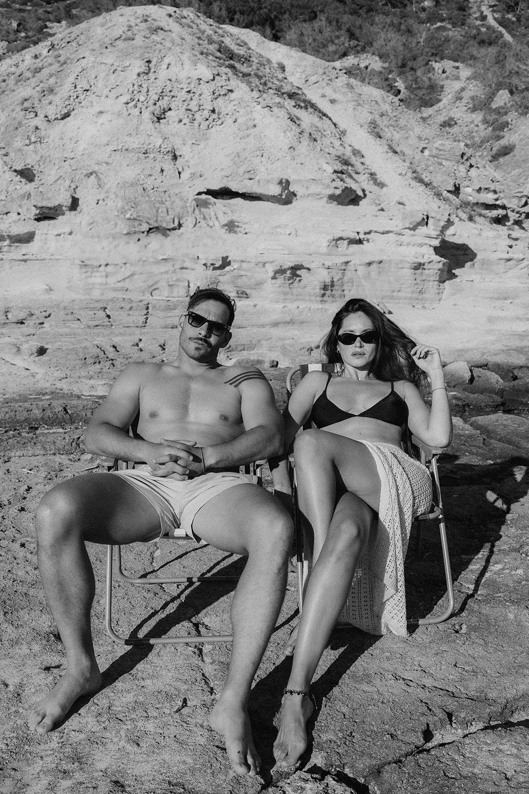 Jimena y Joaquín sentados en sillas de playa frente a una ladera rocosa, en blanco y negro